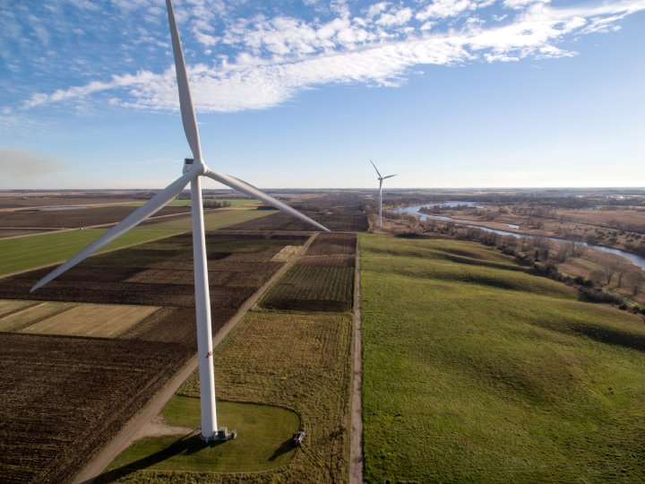 An aerial view of a wind farm in a flat, rural landscape, showing two large white wind turbines standing amidst green fields and brown farmland. A river meanders in the background under a partly cloudy blue sky.